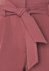 Gros plan d’un pantalon rose poudré avec une ceinture en tissu nouée en nœud à la taille et des coutures visibles sur la poche avant.