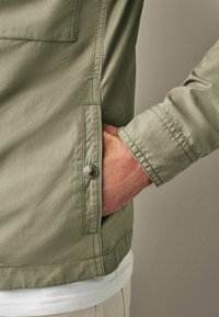 Veste vert olive clair au toucher doux, dotée d'une poche poitrine et d'une fermeture à boutons-pression, présentée avec une main dans la poche latérale.