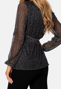 Blouse noire transparente avec détails en fil métallique, manches longues bouffantes et taille péplum plissée. Le tissu texturé renforce l'intérêt visuel.