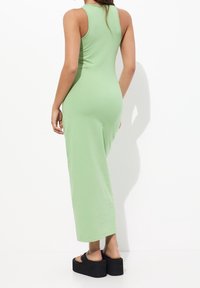 Kobieta w bezrękawowej, jasnzielonej maxi sukience i czarnych sandałach na platformie stoi na tle białym.