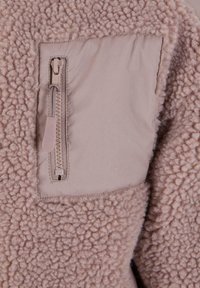 Fleecejakke i myk beige med en glidelås lomme i glatt stoff, med en teksturert, fluffy overflate og kontrasterende panel.