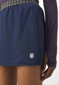 Námořnická modrá sportovní sukně s vzorovaným pasem, s logem K-Swiss. Hladká textura a moderní design vhodný pro sportovní aktivity.