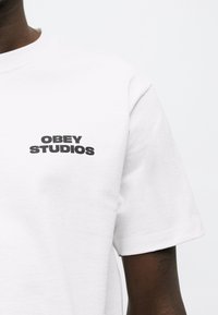 Άτομο που φοράει λευκό κοντομάνικο πουκάμισο με μαύρο κείμενο "OBEY STUDIOS" στο αριστερό στήθος, πάνω σε απλό λευκό φόντο.