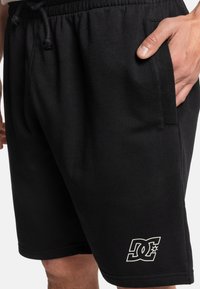 Zwarte katoenen shorts hebben een elastische tailleband met een touwtjesluiting, zijzakken en een wit logo dat op het onderste deel van het linkerpand is gedrukt.