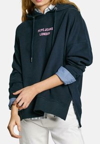 Felpa blu navy con logo rosa "PEPE JEANS LONDON", dotata di cordini e spacchi laterali, indossata sopra una camicia azzurra con colletto.