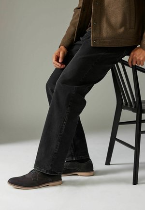 Jeans negros de pierna recta, una chaqueta de punto marrón y zapatos negros con suelas de color claro. El conjunto presenta elementos de diseño simples.
