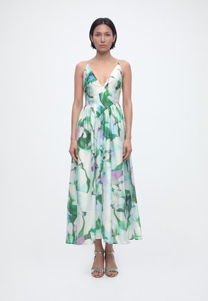 Γυναίκα στέκεται φορώντας μακριά αμάνικη φόρεμα με πράσινο, λευκό και μοβ ανθισμένο μοτίβο και ασημένια ανοιχτά τακούνια, μπροστά σε απλό φόντο.