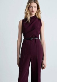 Combinaison sans manches bordeaux avec un décolleté drapé et une ceinture noire ajustée, confectionnée dans un tissu lisse avec une coupe large.