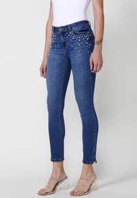 Skinny blue denim jeans featuring pearl embellishments on the front, classic five-pocket design, and hem with rolled cuffs.