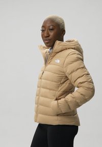 Chaqueta acolchada beige con cierre de cremallera y capucha, que presenta un diseño acolchado y un logotipo blanco en la parte izquierda del pecho. Material sintético suave.