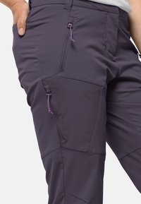 Nahaufnahme von dunkelvioletten Outdoor-Hosen mit schrägen Reißverschlusstaschen und einer Hand, die in der Seitentasche ruht.
