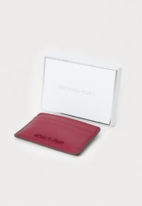 Bordo odinė kortelių laikiklis, ant kurio yra "Michael Kors" logotipas, šalia jo - balta dėžutė su tuo pačiu prekės ženklu.