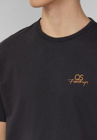 Černé bavlněné tričko s kulatým výstřihem a krátkým rukávem; na levé části hrudníku se nachází vyšitý oranžový logo.