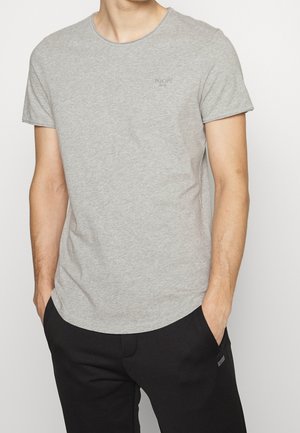 Mężczyzna ubrany w jasnoszary t-shirt z krótkim rękawem i okrągłym dekoltem oraz czarne spodnie, ze rękami w kieszeniach, stojący na jednolitym białym tle.