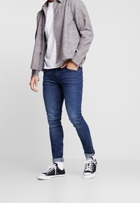 Slim-fit mörkblå jeans, vit t-shirt, grå-rutig skjorta och svarta låga sneakers med vita sulor, stående mot en enfärgad bakgrund.