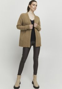 Beige stickad cardigan med öppen framsidan och två fickor. Bärs över en svart spets-topp och kombineras med mörkbruna åtsittande byxor och beige ankelstövlar.