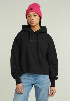 Black cropped hoodie with a front kangaroo pocket and drawstrings, featuring a subtle logo in a textured finish. Paired with light blue jeans.