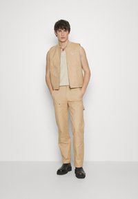 Beige bezramenný svetr přes texturovaný top, doplněný o odpovídající béžové kalhoty a černé kotníkové boty. Jednoduché linie s praktickými kapsami.