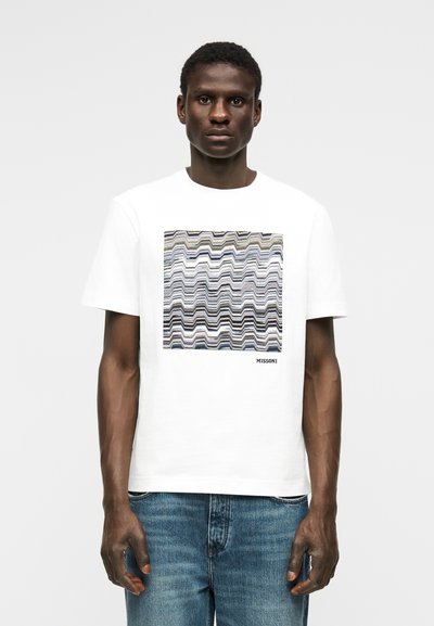 Άνδρας που φορεί λευκό μπλουζάκι με τετράγωνο, πολύχρωμο κυματιστό μοτίβο ρίγες και κείμενο "MISSONI", συνδυασμένο με μπλε τζιν, στέκεται μπροστά σε απλό φόντο.