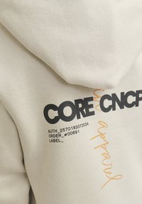 Hoodie od svijetlosmeđe tkanine s teksturiranim finišem. Sadrži otisnuti naslov i podebljani crni tekst s narančastim naglascima.