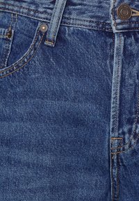 Džínovina ve vybledlé modré barvě, s knoflíkovým zapínáním, měděnými nýtky a klasickým designem s pěti kapsami s žlutými švy.