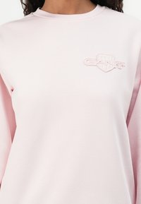 Lichtroze sweatshirt met een ronde hals, lange mouwen en een subtiele geborduurde logo op de borst. Soepele textuur en casual ontwerp.