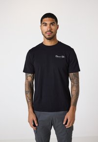 Schwarzes Baumwoll-T-Shirt mit Rundhalsausschnitt und kurzen Ärmeln, verziert mit einem kleinen weißen Logo auf der Brust. Modell trägt graue Cargo-Hosen.