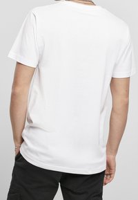 Person som bär en enkel vit T-shirt med kort ärm och svarta byxor, sedd bakifrån, med en silverkedja och armband synliga.
