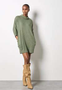 Abito in maglia verde oversize con collo a cowl, dotato di maniche a tre quarti e tasche laterali, abbinato a stivali morbidi beige fino al ginocchio.