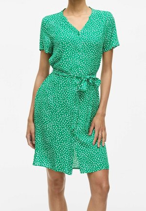 Femme portant une robe verte à manches courtes, longueur au genou, avec des pois blancs, un col en V, des boutons sur le devant et une ceinture nouée à la taille.