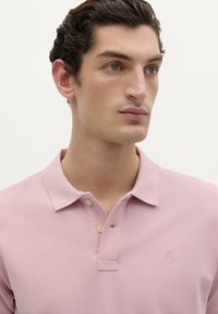 Polo shirt rose clair en tissu texturé, avec une patte de boutonnage à deux boutons et un petit logo brodé sur la poitrine.