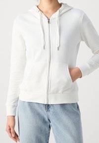 Felpa bianca con zip realizzata in tessuto morbido, dotata di cappuccio, stringhe e tasche frontali. Abbinata a jeans azzurri chiari.