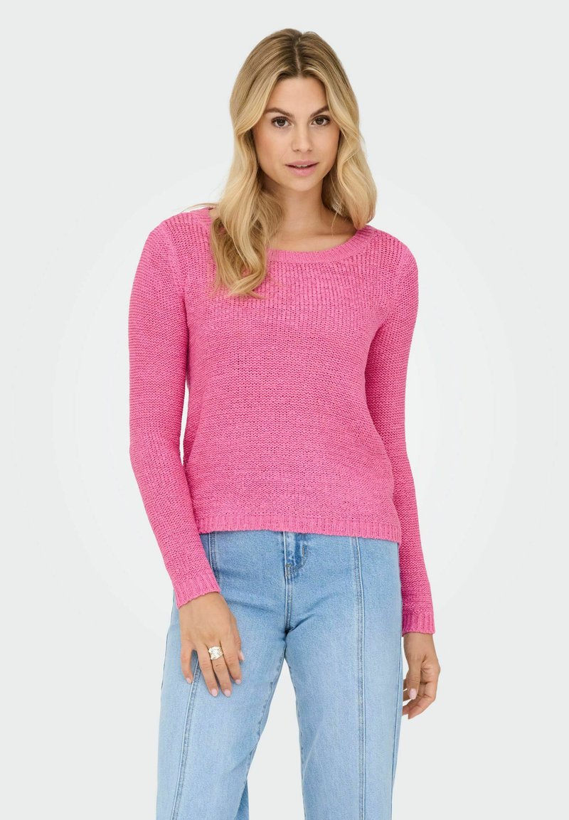 Femme aux longs cheveux blonds portant un pull en tricot rose vif et un jean bleu clair, debout les mains détendues le long du corps.