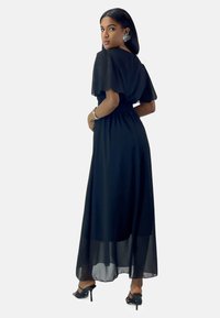 Robe maxi noire avec des manches en éventail transparentes, taille cintrée et jupe fluide. Une paire de sandales à talons hauts achève le look.
