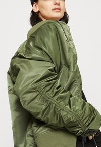 Olive green bomber jacket with a shiny finish, ribbed collar, and elastic cuffs. Features puffy sleeves and visible stitching details.