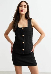 Zwart tweedelig outfit met een getextureerd mouwloos top met vierkante halslijn en gouden knopen, gecombineerd met een aansluitende mini-jurk.