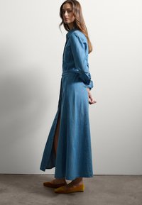 Abito lungo in denim con colletto, vita cintrata e spacchi laterali. Il tessuto è blu con una consistenza morbida. Indossato con scarpe ballerine gialle.