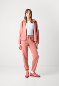 Vrouw in een roze zip-up hoodie, bijpassende sweatpants, wit topje en roze sneakers, staande tegen een egale witte achtergrond.