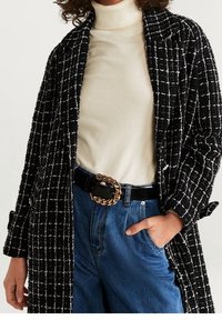 Manteau texturé noir et blanc à motif à carreaux, col roulé beige, jean bleu taille haute, et ceinture noire avec boucle chaîne dorée.