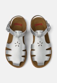 Hopeakupuiset sandaalit, joissa on aukkoaukot ja solkiketju. Ruskea pohja, jossa on pyöreitä reikiä ilmanvaihdon takaamiseksi. Brandin nimi "CAMPER" on painettu punaisella.