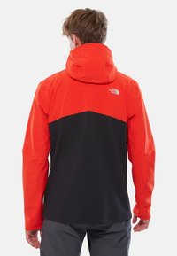 Röd och svart vattentät jacka med huva, som har en färgblockdesign och logodetalj på ryggen. Tillverkad av ett mjukt, andningsbart material.