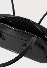 Crna kožna torba s zlatnim zatvaračem, dvostrukim ručkama i teksturiranim završetkom. Unutrašnjost uključuje džep sa zatvaračem i prostrani glavni odjeljak.