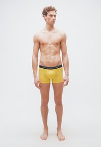 Νέος άνδρας στέκεται ξυπόλητος φορώντας κιτρινόξανθα εσώρουχα Polo Ralph Lauren μπροστά σε ένα απλό λευκό φόντο.