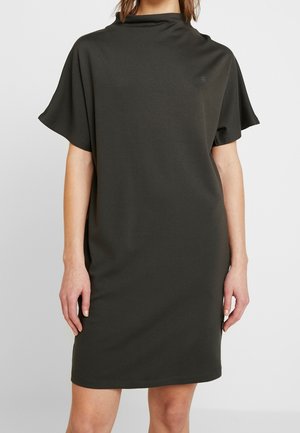 Kvinde iført en løs, kortærmet mørkegrå kjole i knælængde med en høj, bred halsudskæring, stående foran en ensfarvet lys baggrund.