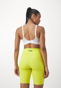 Witte sportbeha en felgele high-waisted fietsbroek. De stof is glad; de broek heeft een getailleerd ontwerp en een achterzak.