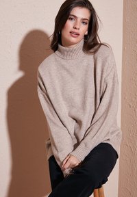 Bež turtleneck-sviiter pehmest tekstureeritud kangast; lahtine lõige ja soonikkoes mansetid. Kandub mustade pükste kombineerituna, istudes puidust toolil.