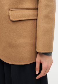 Κοντινό πλάνο του χεριού ενός ατόμου δίπλα σε παλτό από μαλλί σε χρώμα καμήλας με τσέπη με καπάκι, και μαύρο παντελόνι ορατό από κάτω.