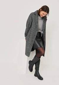 Szürke texturaú kabát, amely gall�érral rendelkezik, egy világosszürke pulóver és fekete bőrszoknya fölé rétegezve, térd fölé érő fekete csizmával párosítva.