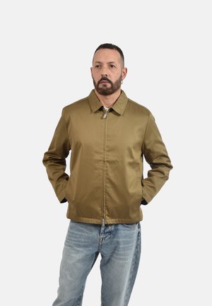 Uomo con capelli corti e barba indossa una giacca marrone con zip e jeans azzurri, in piedi con le mani nelle tasche su sfondo uniforme.