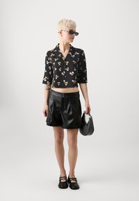 Svart blomstret cropped skjorte med korte ermer, kombinert med svarte faux skinnshorts med folder og to frontknapper. Svart håndveske og sko.